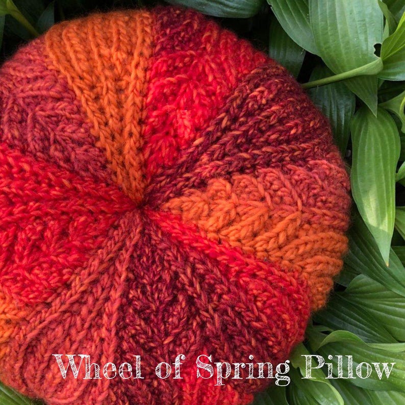 May include: A round, crocheted cushion with a colourful, ombre design in shades of red, orange, and brown. The cushion is labelled "Wheel of Spring Pillow".