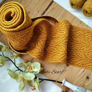 Puede incluir: Una bufanda de punto de color amarillo dorado con un patr&oacute;n texturizado. La bufanda est&aacute; enrollada y se muestra sobre una superficie de madera con un arreglo floral.