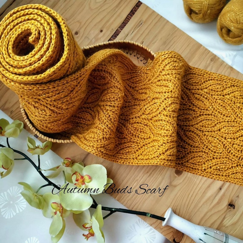 Puede incluir: Una bufanda de punto de color amarillo dorado con un patr&oacute;n texturizado. La bufanda est&aacute; enrollada y se muestra sobre una superficie de madera con un arreglo floral.