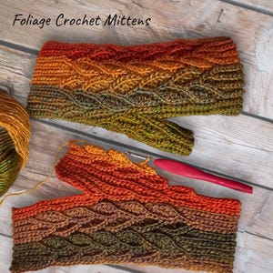 Peut inclure: Une paire de mitaines en crochet sans doigts dans des tons de vert, orange et marron. Les mitaines présentent un motif de tricot torsadé et sont réalisées avec un fil chiné. Le texte "Foliage Crochet Mittens" est visible sur l'image.