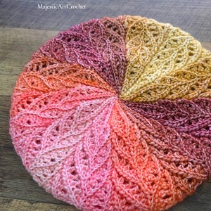 May include: A round crochet beret featuring a radial leaf pattern in a gradient of colors including yellow, orange, pink, and deep purple. The text Clematis Beret and MajesticArtCrochet is visible at the top of the image.
