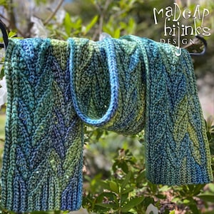 May include: A blue and green knitted scarf with a cable knit pattern. The scarf is hanging on a metal hook. The text "Madcap Hijinks Design" is visible in the top right corner of the image.