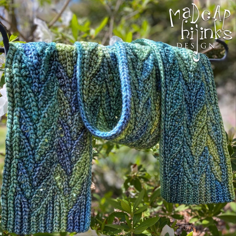 May include: A blue and green knitted scarf with a cable knit pattern. The scarf is hanging on a metal hook. The text "Madcap Hijinks Design" is visible in the top right corner of the image.