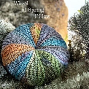 May include: A colourful crocheted pillow with a wheel of spring design. The pillow is made with a variety of shades of blue, green, purple, and orange yarn.