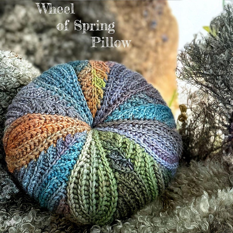 May include: A colourful crocheted pillow with a wheel of spring design. The pillow is made with a variety of shades of blue, green, purple, and orange yarn.