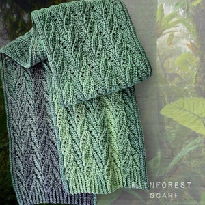 May include: Two crocheted scarves in a green and blue ombre pattern. The scarves are folded and laid on a green background with the text "RAINFOREST SCARF".