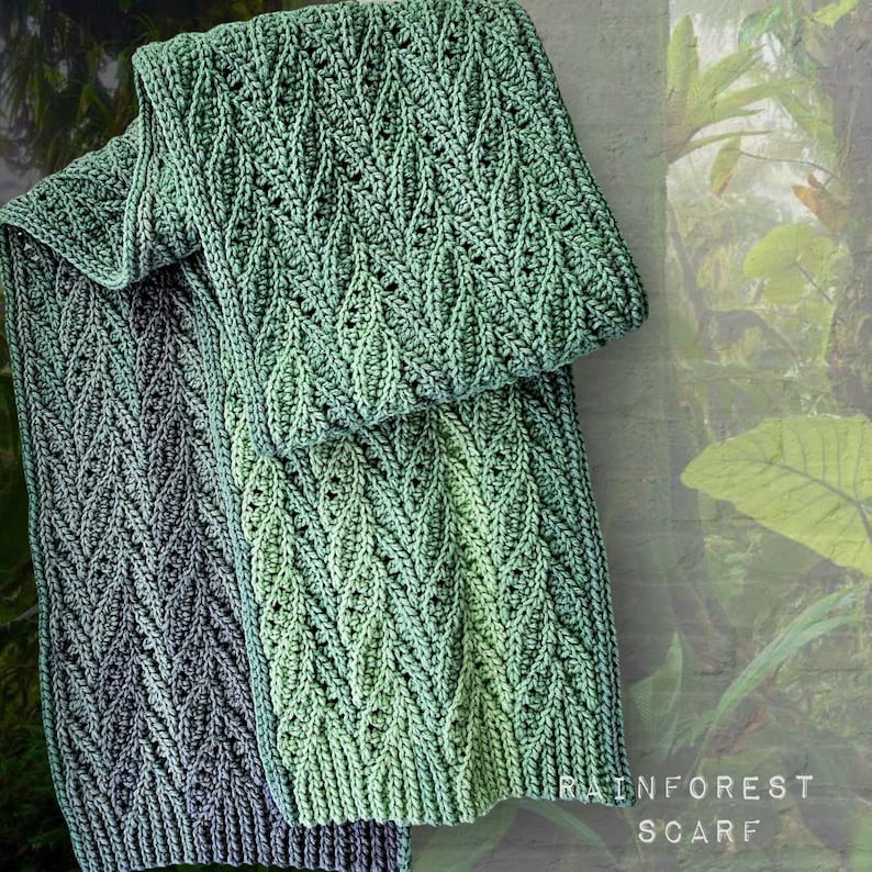 May include: Two crocheted scarves in a green and blue ombre pattern. The scarves are folded and laid on a green background with the text "RAINFOREST SCARF".