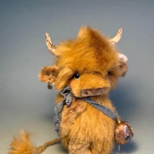 May include: A handcrafted plush Highland cow toy with light brown, shaggy fur. It features small, curved horns, a gray scarf, and a bell. The toy cow has a fluffy tail and a detailed, artisan appearance.