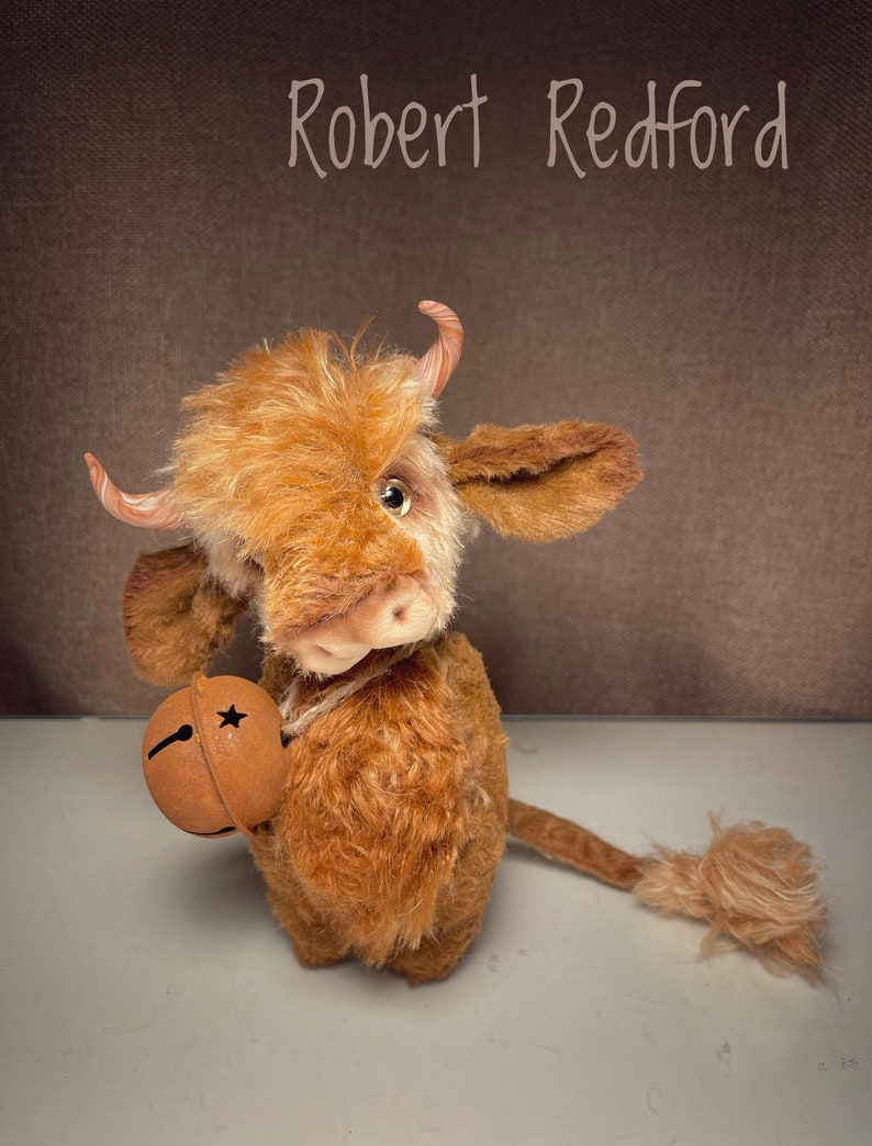 May include: A brown and white plush toy cow with a bell in its hand. The cow has a long tail and is standing on a white surface. The text "Robert Redford" is written above the cow.