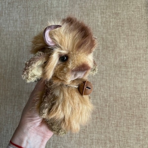 May include: A small, brown and white plush toy bison with a pink horn and a bell around its neck.
