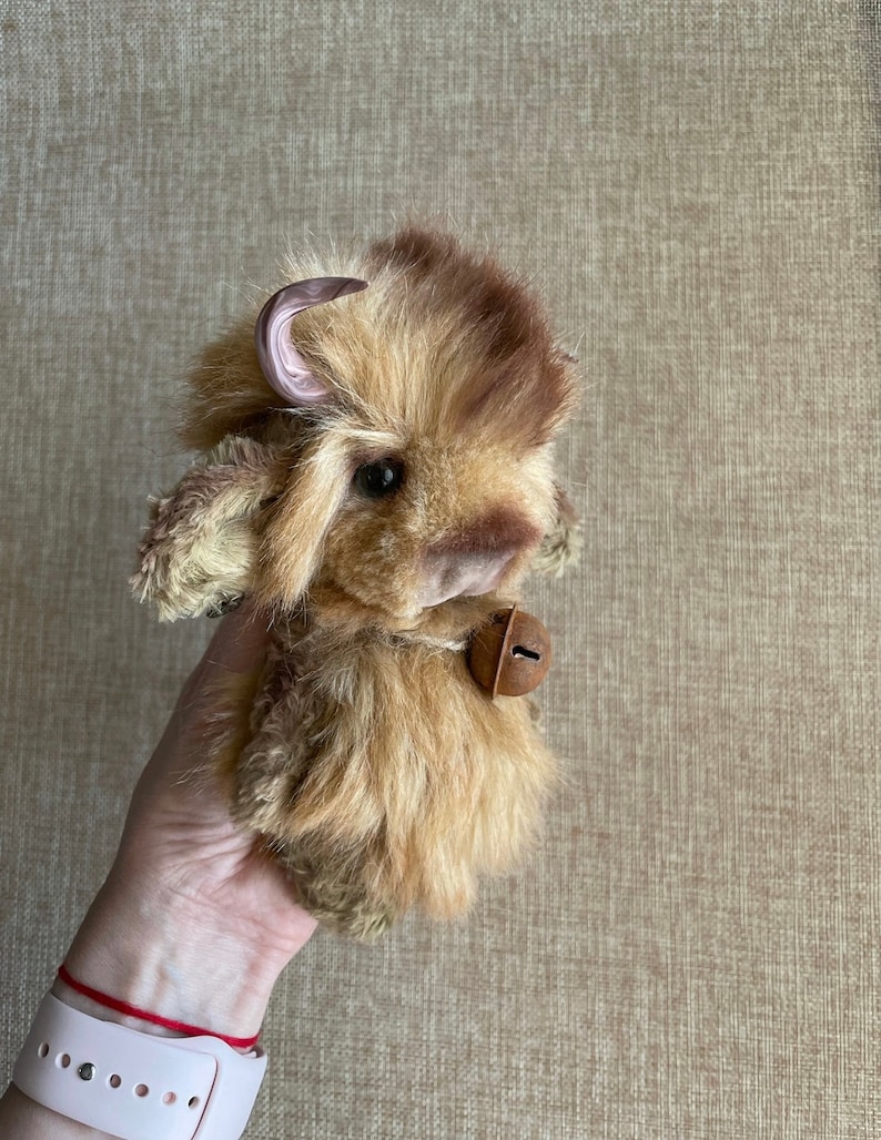 May include: A small, brown and white plush toy bison with a pink horn and a bell around its neck.