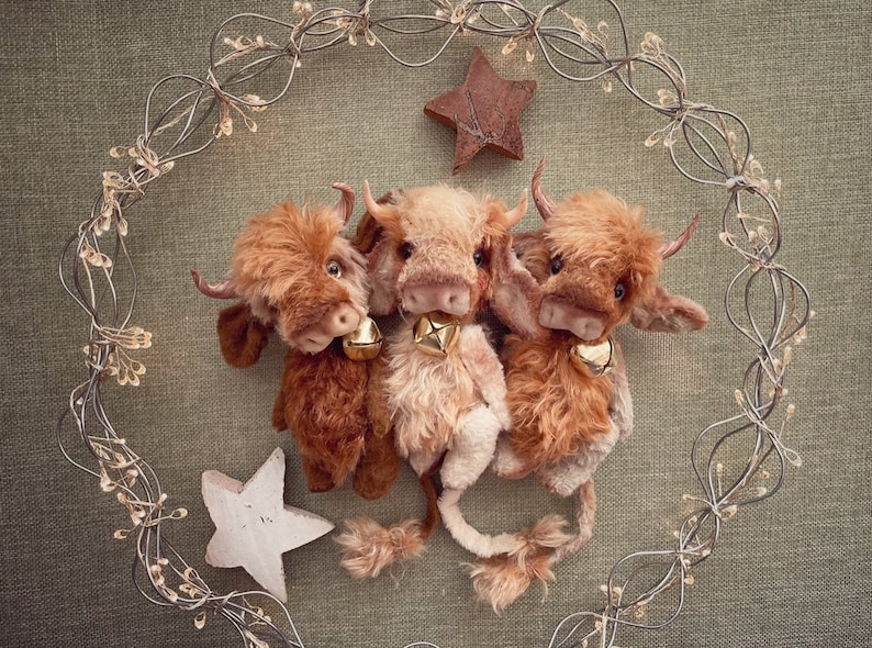 May include: Three brown and white plush toy Highland cows with bells around their necks are arranged in a circle against a green fabric background. A wire wreath with lights is behind the toys.