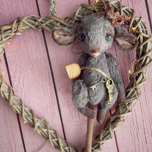 Puede incluir: Un ratón de peluche gris con una llave y un trozo de queso colgando de su cuello. El ratón está sentado en una rama marrón dentro de una corona de mimbre en forma de corazón.