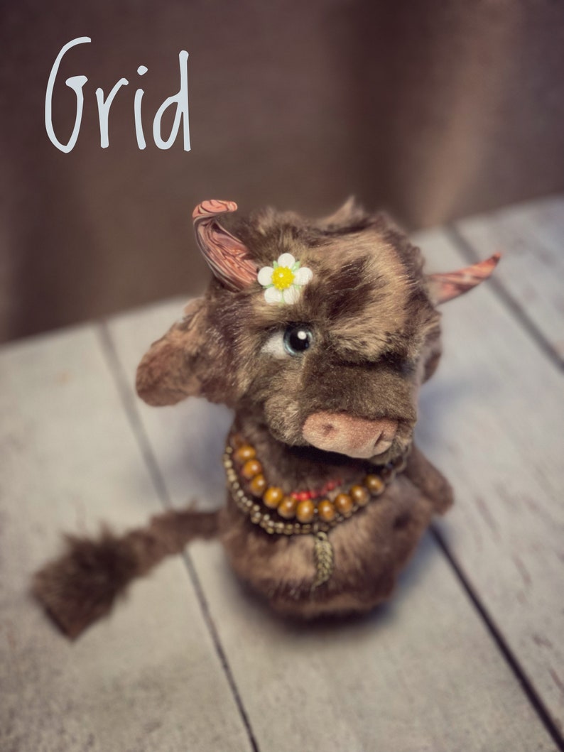 May include: A brown and black plush toy cow with a white flower on its head. The cow has a beaded necklace and a long tail. The word "Grid" is written in white on a brown background.