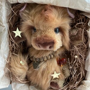 May include: A brown and tan plush toy with a long, fluffy tail. The toy has a beaded necklace and a small, brown heart charm. The toy is sitting in a nest of shredded paper with gold star-shaped confetti.