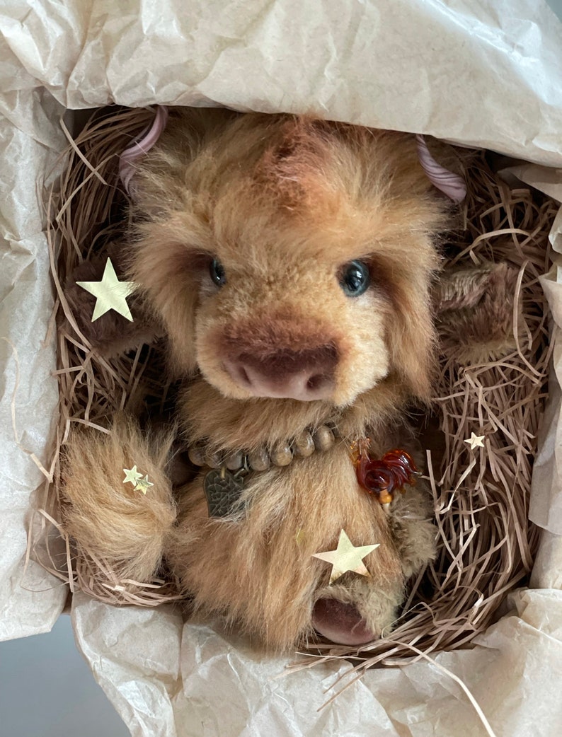May include: A brown and tan plush toy with a long, fluffy tail. The toy has a beaded necklace and a small, brown heart charm. The toy is sitting in a nest of shredded paper with gold star-shaped confetti.