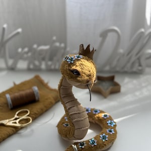 Può includere: Un serpente di peluche fatto a mano con una corona e decorazioni floreali. Il serpente è principalmente marrone con dettagli beige e blu. Ha una piccola corona e un anello d'argento sulla coda. Il serpente è in posizione eretta.