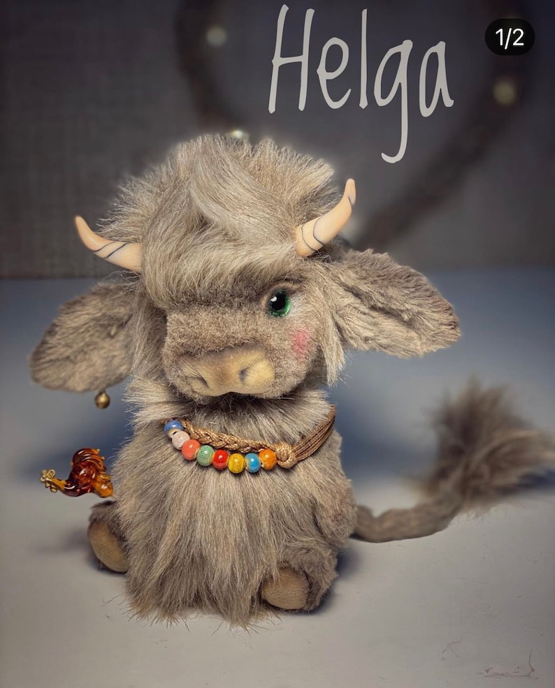 May include: A gray and brown plush toy yak with a long tail, horns, and a colorful beaded necklace. The toy is sitting on a light gray surface. The text "Helga 1/2" is visible in the upper right corner of the image.