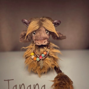 May include: A brown and tan plush toy with a long tail and a beaded necklace. The toy has a long, shaggy mane and a long, shaggy tail. The toy is sitting on a white surface and the text "Tanana" is written below the toy.