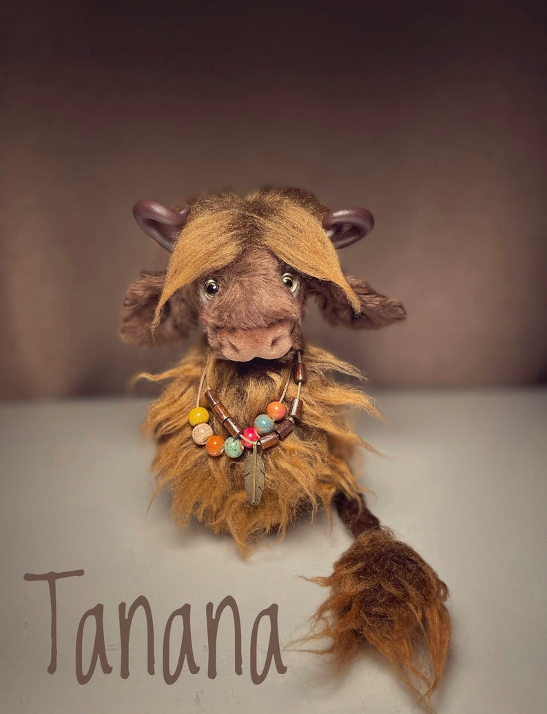 May include: A brown and tan plush toy with a long tail and a beaded necklace. The toy has a long, shaggy mane and a long, shaggy tail. The toy is sitting on a white surface and the text "Tanana" is written below the toy.