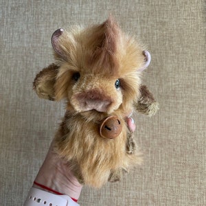 May include: A brown and tan plush toy cow with a bell around its neck. The cow has a fluffy brown and tan coat and a small, brown nose.
