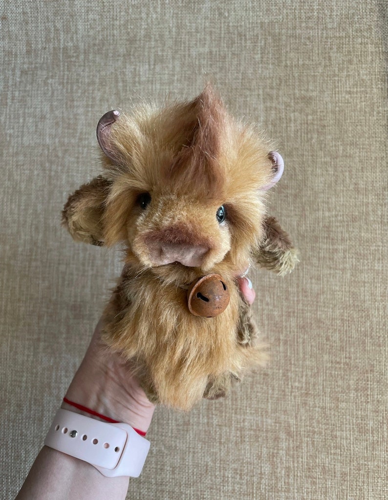 May include: A brown and tan plush toy cow with a bell around its neck. The cow has a fluffy brown and tan coat and a small, brown nose.