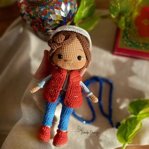 May include: A crocheted doll wearing a red vest, blue jeans, and a white hat. The doll has brown hair and green eyes. The doll is holding a blue beaded necklace.