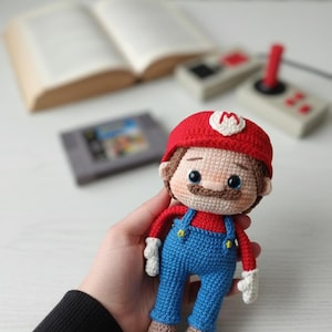 Puede incluir: Muñeco de Mario tejido a ganchillo, con gorro rojo con una 'M' blanca, peto azul y zapatos marrones. El muñeco tiene bigote marrón y sostiene un mando y un cartucho de juego. Un libro y un mando de juego retro en el fondo.