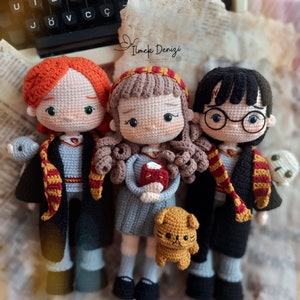 May include: Three crocheted dolls, two girls and one boy, dressed in Hogwarts school uniforms. The girl on the left has red hair and is holding a small crocheted owl. The girl in the middle has brown hair and is holding a book. The boy on the right has black hair and is wearing glasses. He is holding a small crocheted ghost. A small crocheted cat is in front of the dolls.