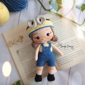 May include: A crocheted doll with brown hair, wearing a yellow and blue hat with goggles, a blue overall, and black shoes. The doll is sitting on a book with the text "Ilmek Denizi" on the cover.