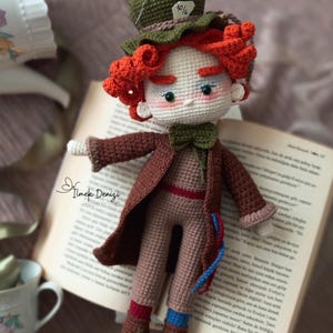 Könnte beinhalten: Eine gehäkelte Puppe des Hutmachers aus Alice im Wunderland. Die Puppe trägt einen braunen Mantel, eine braune Hose, einen grünen Hut mit dem Text "10/6" darauf und rote Haare. Die Puppe sitzt auf einem Buch.
