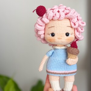 以下が含まれることがあります： ピンクの縮れた髪、青いドレス、茶色の靴を履いた、かぎ針編みのドール。ドールは、赤いチェリーが乗った、かぎ針編みのアイスクリームコーンを持っています。