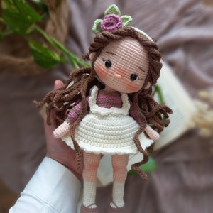 May include: A crocheted doll with brown hair, a pink flower crown, and a white dress. The doll is wearing pink and white striped sleeves and brown shoes.