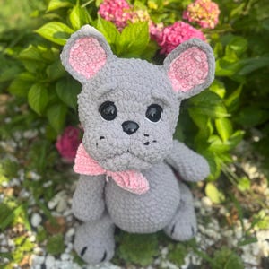May include: A handmade gray crochet mouse with pink inner ears and a pink bow. The plush toy has black eyes and a black nose. The mouse is sitting in front of green foliage and pink flowers.