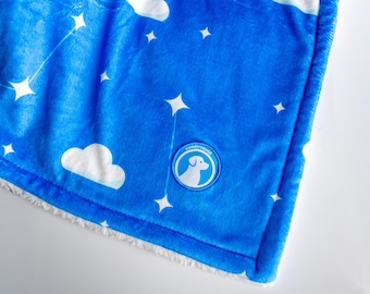 May include: A blue plush blanket with a constellation and cloud pattern. White stars and clouds are scattered across the blanket. The blanket has a white, fluffy border and a circular logo that says "wondersquad" with a dog silhouette.