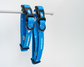 May include: Two blue dog collars with black buckles and D-rings hang from a white bar. The collars have a pattern of white clouds and stars. The text "Wondersquad" is printed on the collar.