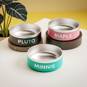 May include: Three stainless steel pet bowls with wooden stands. The bowls are labeled "Pluto", "Maple", and "Minnie".
