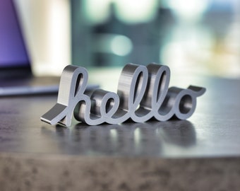 May include: A silver metal word "hello" in a cursive font. The word is 3D and stands on a gray surface.