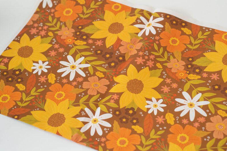 May include: A roll of floral patterned paper featuring a repeating design of sunflowers, daisies, and other flowers in shades of yellow, orange, and white on a brown background. The paper is partially unrolled.