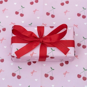 May include: A rectangular gift box wrapped in pink paper with a cherry pattern and tied with a red satin ribbon. The background is a matching pink paper with the same cherry design.