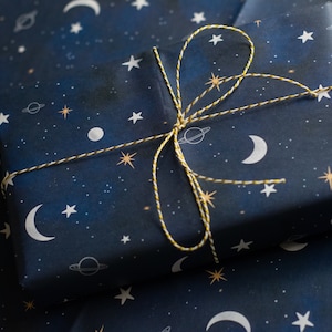 May include: A wrapped gift box featuring dark blue paper with a celestial pattern of white moons, stars, planets, and dots. The box is tied with a gold and white twine bow, creating a festive presentation. Perfect for birthdays or special occasions.
