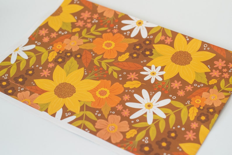 May include: A sheet of paper with a repeating floral pattern. The design features large yellow sunflowers, orange and white flowers, and green leaves on a brown background. The overall theme is autumnal.
