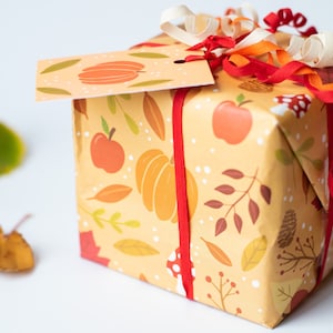 May include: A gift box wrapped in fall-themed paper with a red ribbon and a gift tag that says "Happy Fall". The paper features pumpkins, apples, leaves, and branches in shades of orange, red, and brown.