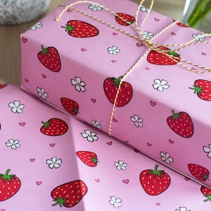 May include: Pink gift wrap with a repeating pattern of strawberries, white flowers, and pink hearts. The gift wrap is tied with a gold string.