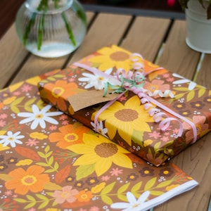 May include: A wrapped gift with a brown floral pattern featuring sunflowers, daisies, and orange flowers. The gift is tied with pink ribbon and a gift tag. A matching sheet of wrapping paper is below.