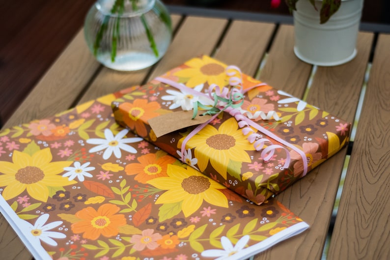 May include: A wrapped gift with a brown floral pattern featuring sunflowers, daisies, and orange flowers. The gift is tied with pink ribbon and a gift tag. A matching sheet of wrapping paper is below.