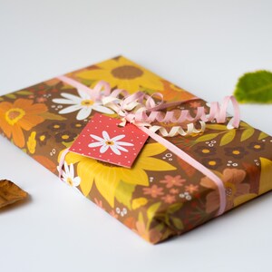 May include: A wrapped gift with floral patterned paper in shades of brown, yellow, and orange. A pink ribbon and gift tag with a white daisy design adorn the package. Dried leaves are scattered around the gift.
