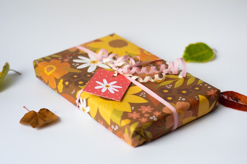 May include: A wrapped gift with floral patterned paper in shades of brown, yellow, and orange. A pink ribbon and gift tag with a white daisy design adorn the package. Dried leaves are scattered around the gift.