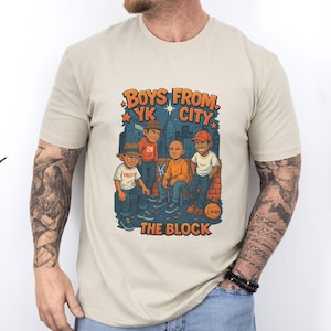 May include: A beige t-shirt featuring a graphic of five cartoon figures with the text "BOYS FROM YK CITY THE BLOCK" in orange and blue. The figures are depicted in various outfits, with a cityscape backdrop.