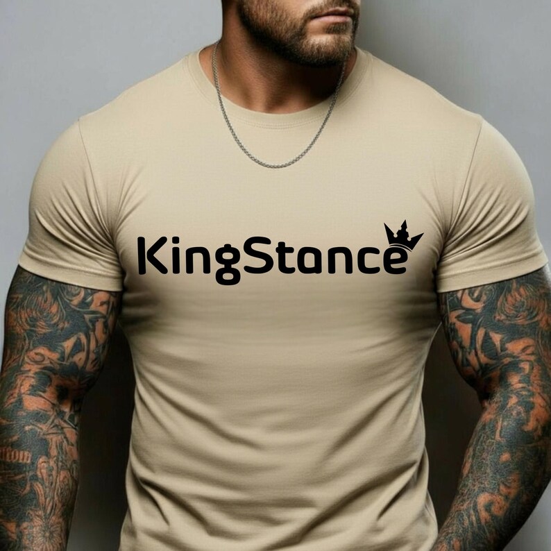 Puede incluir: Una camiseta beige de cuello redondo con el texto negro "KingStance" y un gr&aacute;fico de corona. La camiseta de manga corta est&aacute; hecha de un material suave. El modelo lleva una cadena plateada.